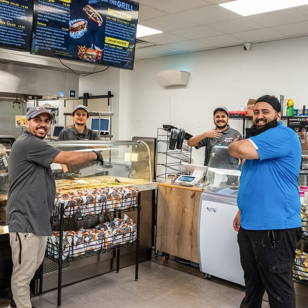 Three smiling male employees stand behind the counter of a deli, with food displays and menus visible.