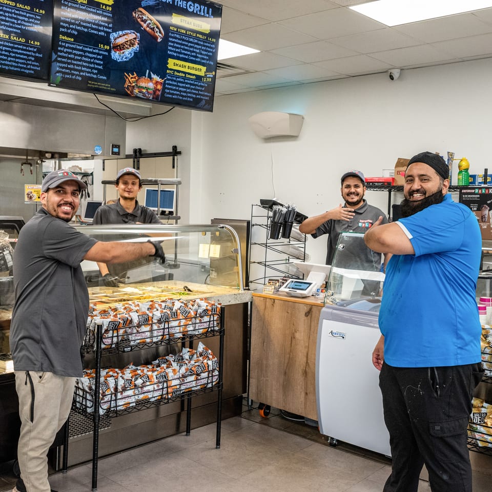 Three smiling male employees stand behind the counter of a deli, with food displays and menus visible.