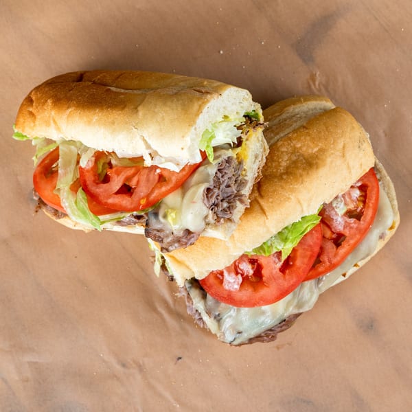 Two halves of a steak and cheese sub sandwich with lettuce and tomato on brown paper.