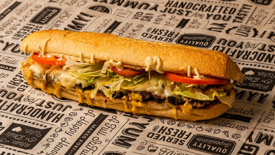 A large submarine sandwich with melted cheese, lettuce, tomatoes, and sauces on patterned paper.
