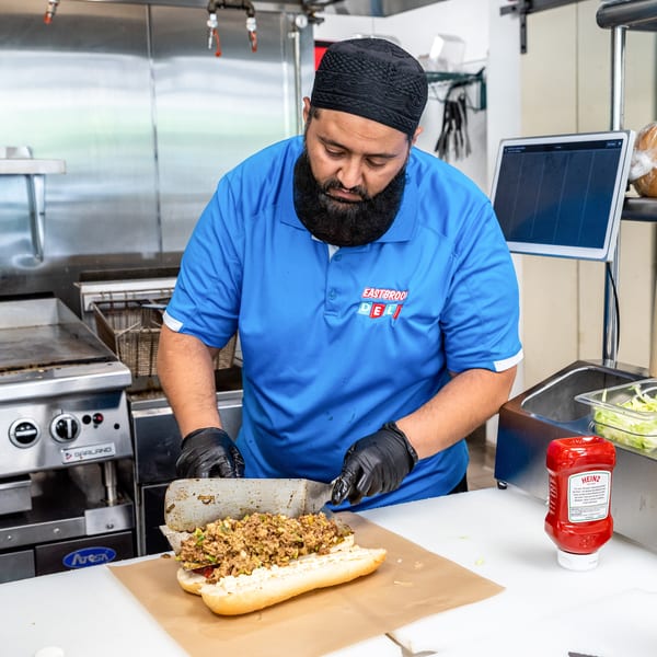 A chef in a blue Eastbrook Deli shirt prepares a cheesesteak sandwich in a commercial kitchen.
