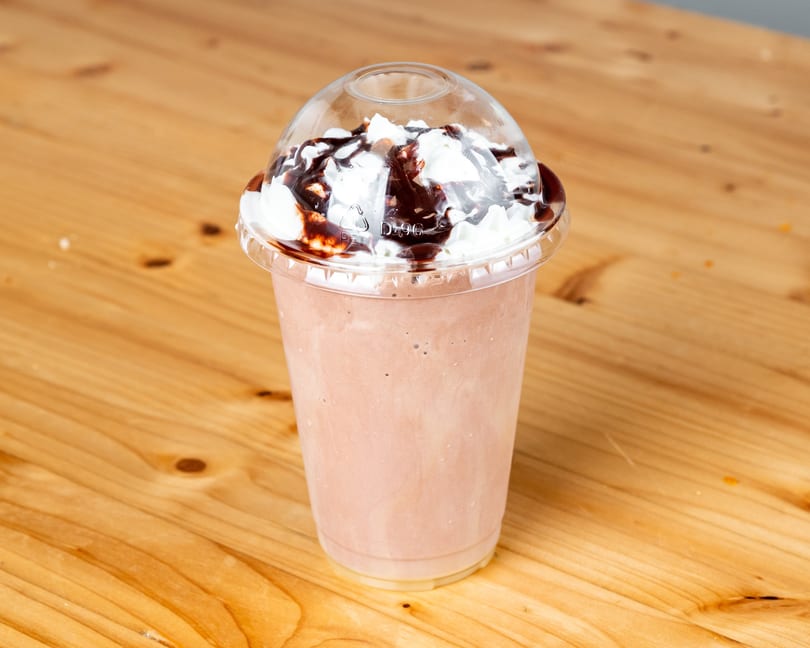Chocolate milkshake with whipped cream and chocolate syrup in a plastic cup on a wooden table.