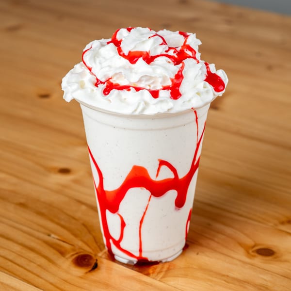 Vanilla milkshake with whipped cream and strawberry syrup drizzle on a wooden table.