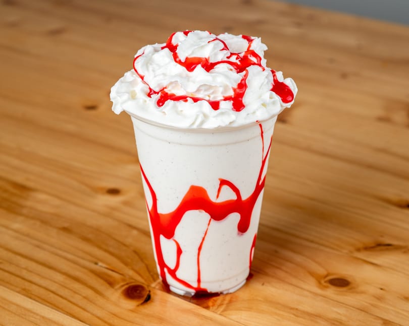 Vanilla milkshake with whipped cream and strawberry syrup drizzle on a wooden table.