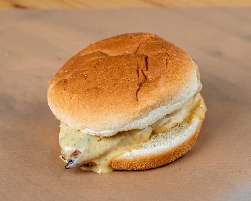 A grilled chicken sandwich with melted cheese and mustard on a bun, on a brown paper surface.