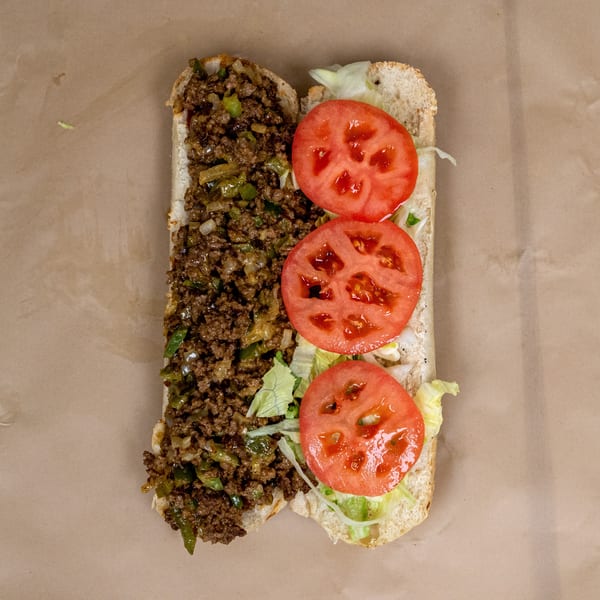 Open-faced cheesesteak with ground meat, peppers, onions, lettuce, and sliced tomatoes on a sub roll.