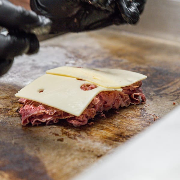 Chef's gloved hands preparing a sandwich with sliced meat and cheese on a griddle.