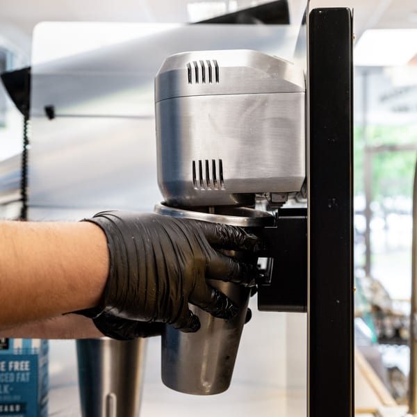 A gloved hand holds a metal cup under a milkshake machine, with a milk carton nearby.