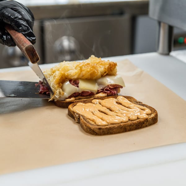 A chef's gloved hand uses a spatula to assemble a hot pastrami sandwich with melted cheese.
