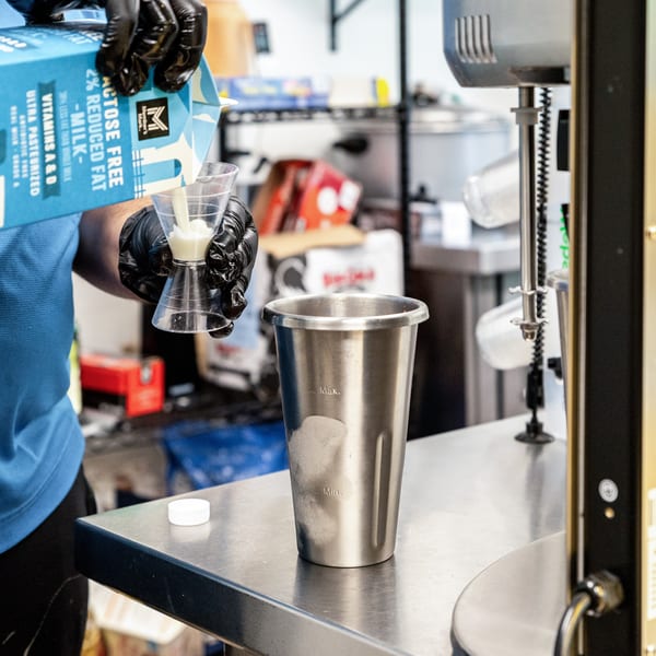 Gloved hands pour milk from a carton into a measuring cup over a metal mixing cup.