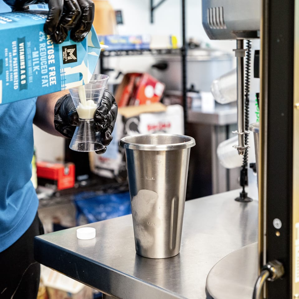 Gloved hands pour milk from a carton into a measuring cup over a metal mixing cup.