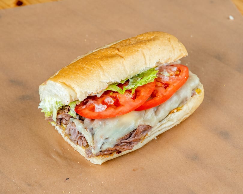 Roast beef sub sandwich with melted cheese, lettuce, and sliced tomatoes on a brown paper.