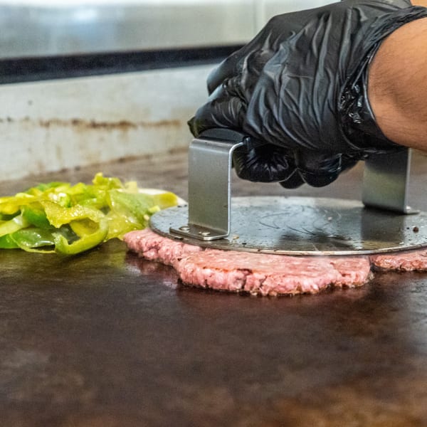 Gloved hand pressing raw burger patties on a griddle next to green peppers.