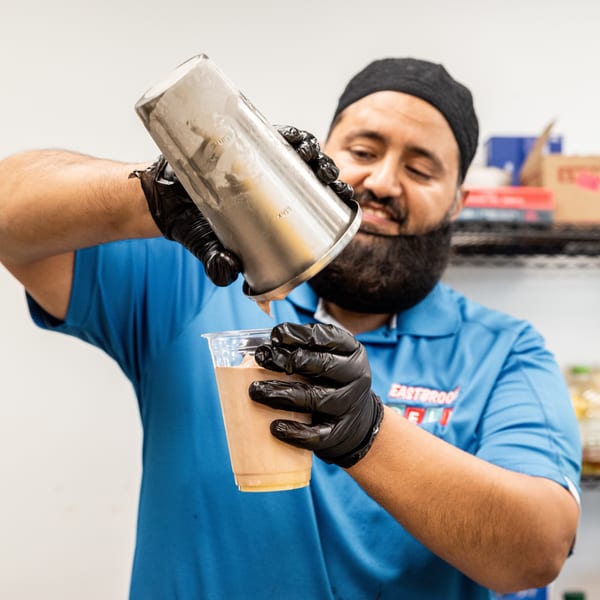 A man in a blue shirt and black gloves pours a drink from a metal shaker into a plastic cup.