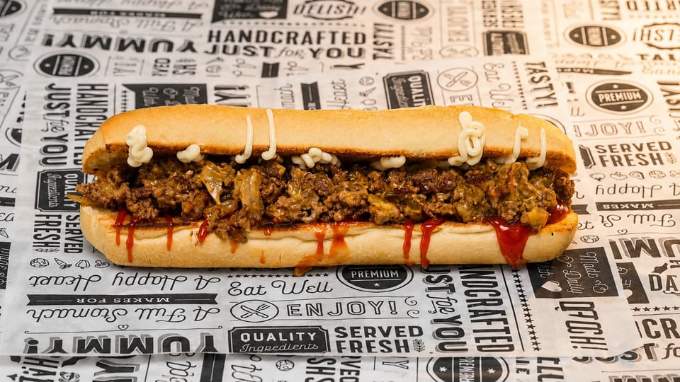 A close-up of a loaded cheesesteak sandwich with sauce on branded deli paper.