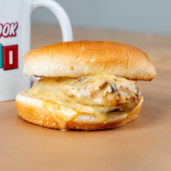 A chicken sandwich with melted cheese and sauce on a bun, next to a white mug.