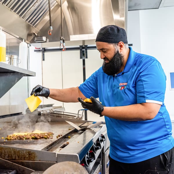A chef in a blue shirt and black gloves adds cheese to sizzling meat and vegetables on a griddle.