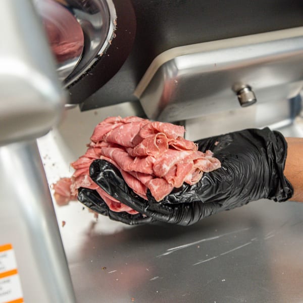 A gloved hand holds a pile of freshly sliced deli meat next to a commercial meat slicer.